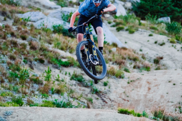 A young cyclist in a helmet and gloves is performing a jump on a mountain bike over rugged terrain, surrounded by rocky landscapes and lush greenery at sunset. Anthony Lakes Mountain Resort mountain bike trail.