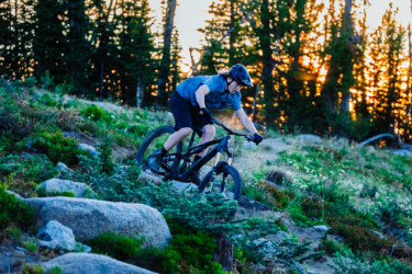 A person riding a mountain bike down a rocky trail surrounded by lush greenery and trees at sunset. The rider is wearing a helmet and sport attire, focused on navigating the terrain. Soft golden light filters through the trees in the background. Anthony Lakes Mountain Resort mountain bike trail.