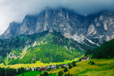 A scenic view of a mountainous landscape featuring towering cliffs and lush green hills, with a small village nestled at the base surrounded by dense forests. The sky is partly cloudy, with fog enveloping the higher peaks. Sellaronda MTB Loop mountain bike trail.