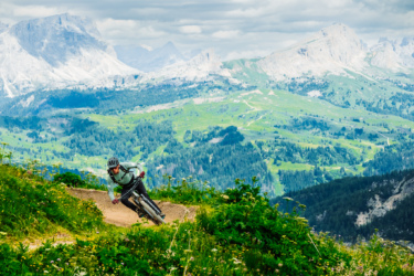 A mountain biker navigating a curved dirt trail on a lush hillside, with mountainous terrain and green valleys in the background under a partly cloudy sky.