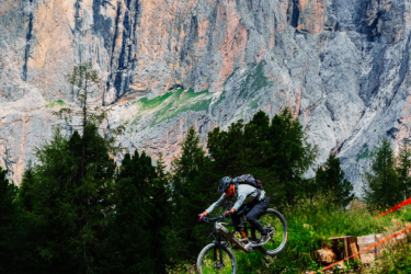 A mountain biker performing a jump on a downhill trail, surrounded by lush greenery and forest, with dramatic rocky mountains in the background under a partly cloudy sky. Red markers outline the trail, indicating a biking course.
