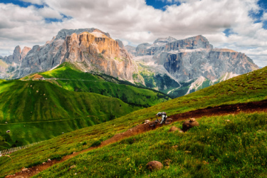 A mountain biker navigates a winding dirt trail through lush green hills, with dramatic rocky peaks and a partly cloudy sky in the background. Sellaronda MTB Loop mountain bike trail.