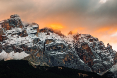 A dramatic mountain landscape at sunset featuring rugged peaks partially covered in snow, with clouds lingering around the summit and vibrant orange hues glowing in the sky. Sellaronda MTB Loop mountain bike trail.