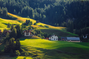 A picturesque landscape featuring rolling green hills illuminated by sunlight, with scattered houses nestled among trees. The background is filled with dense forests, creating a serene and tranquil rural scene. Sellaronda MTB Loop mountain bike trail.