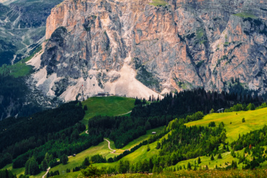 A picturesque landscape featuring a towering rock formation surrounded by lush green hills and forests. A winding dirt path leads through the foreground, gently guiding the viewer's eye toward the dramatic mountain peaks in the background, set against a moody sky filled with clouds. Sellaronda MTB Loop mountain bike trail.