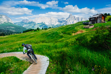 A mountain biker navigating a winding wooden pathway through a lush green landscape, with majestic mountains in the background and a rustic cabin nearby under a bright blue sky dotted with clouds.