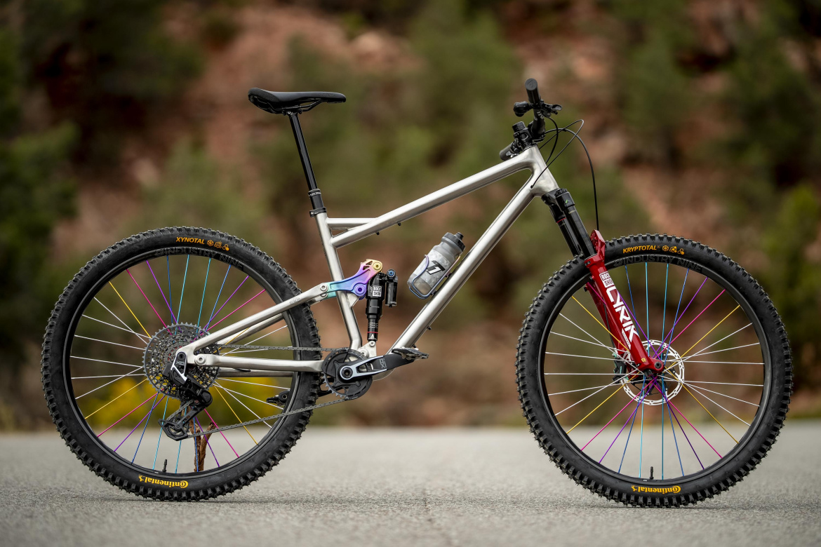 A modern mountain bike featuring a silver aluminum frame, with multicolored spokes on the wheels. The bike has a red front fork, a shock absorber, and a water bottle mounted on the frame. The background shows a blurred natural landscape of greenery and rocks, indicating an outdoor setting.