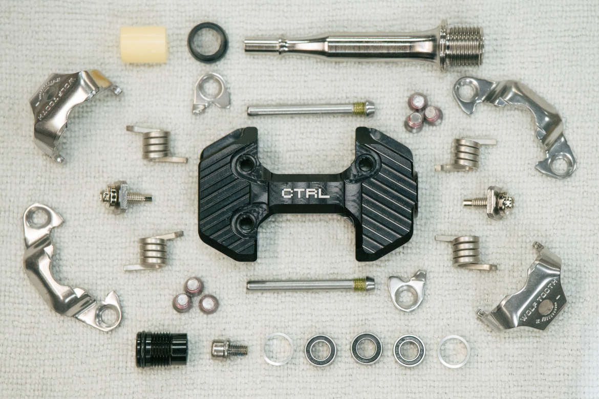 A flat lay of various mechanical components arranged on a textured surface. The center features a black part with the text "CTRL," surrounded by metallic pieces, springs, small screws, and washers, indicating a collection of bike or machinery parts for repair or assembly.
