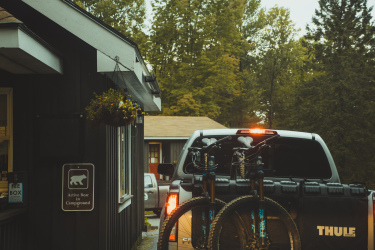 Photo of a pickup truck parked in front of a rustic campground office, featuring two mountain bikes mounted on the truck's bike rack. A sign indicating "Active Bear in Campground" is visible on the building, surrounded by greenery and trees in the background. Perry Hill mountain bike trail.
