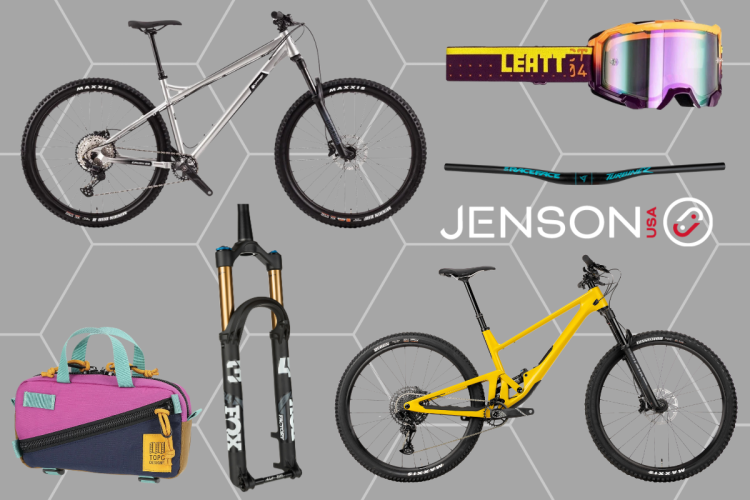 An assortment of mountain biking gear and accessories arranged on a gray background, featuring a silver and a yellow mountain bike, a pair of goggles with a reflective lens, a handlebar, a fork, and a colorful tool bag. The Jenson USA logo is visible in the bottom right corner.