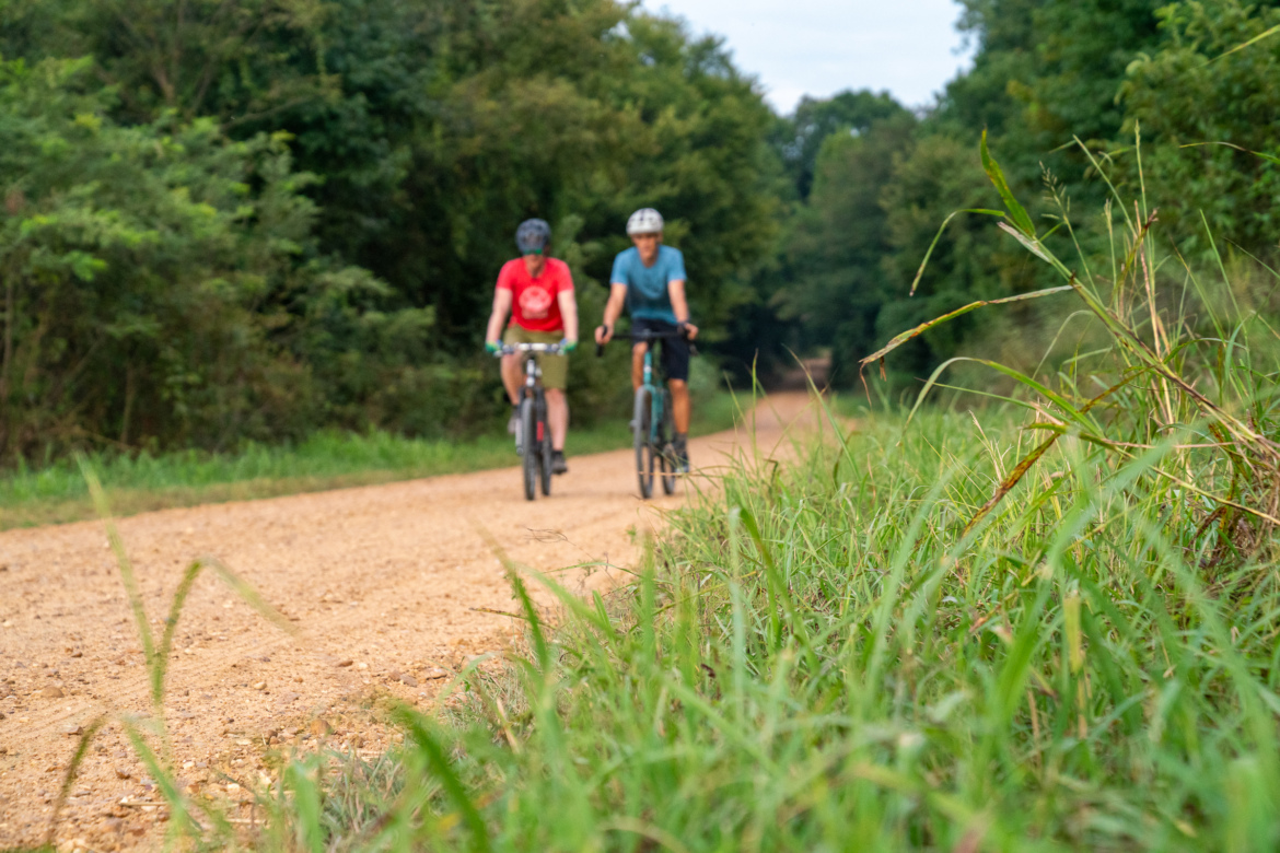 Two cyclists riding on a dirt path surrounded by greenery. One cyclist is wearing a red shirt and shorts, while the other is dressed in a blue shirt. Tall grass is visible in the foreground, adding to the natural scenery.