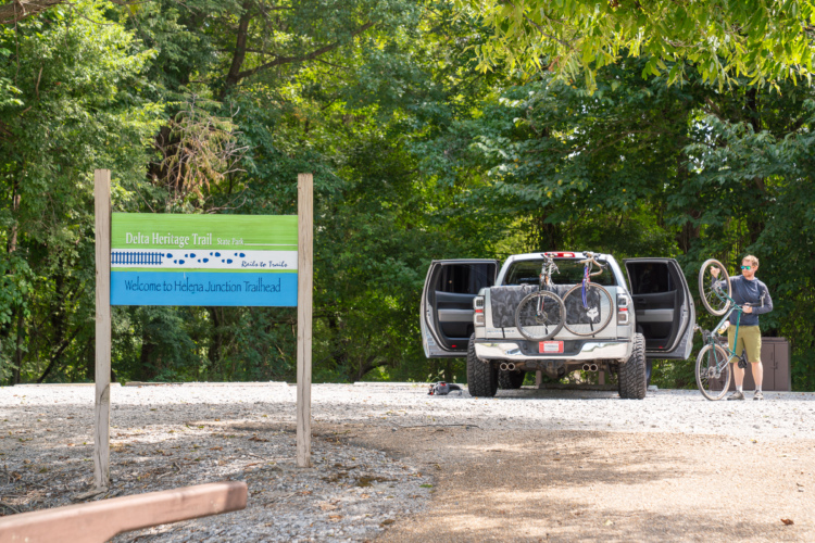 A man prepares his bicycle next to a pickup truck with open doors, parked at the Helena Junction Trailhead. A sign reads "Delta Heritage Trail State Park - Welcome to Helena Junction Trailhead," surrounded by lush green trees.