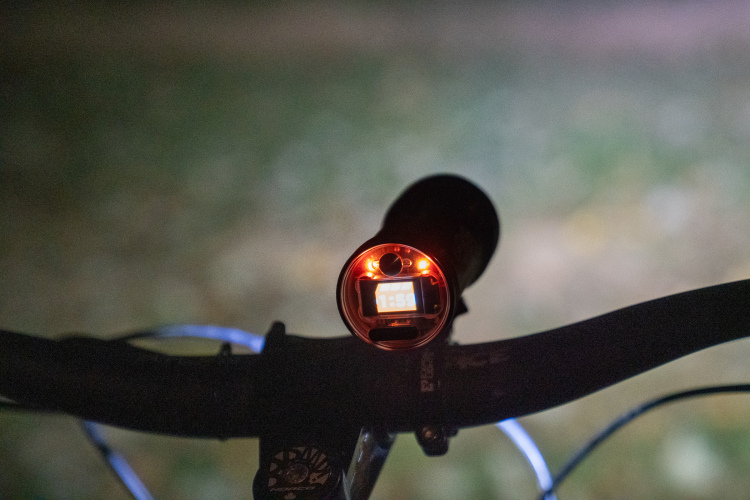 Close-up view of a bicycle handlebar with a mounted front light displaying a digital timer. The light emits a faint orange glow against a dark, blurred background.