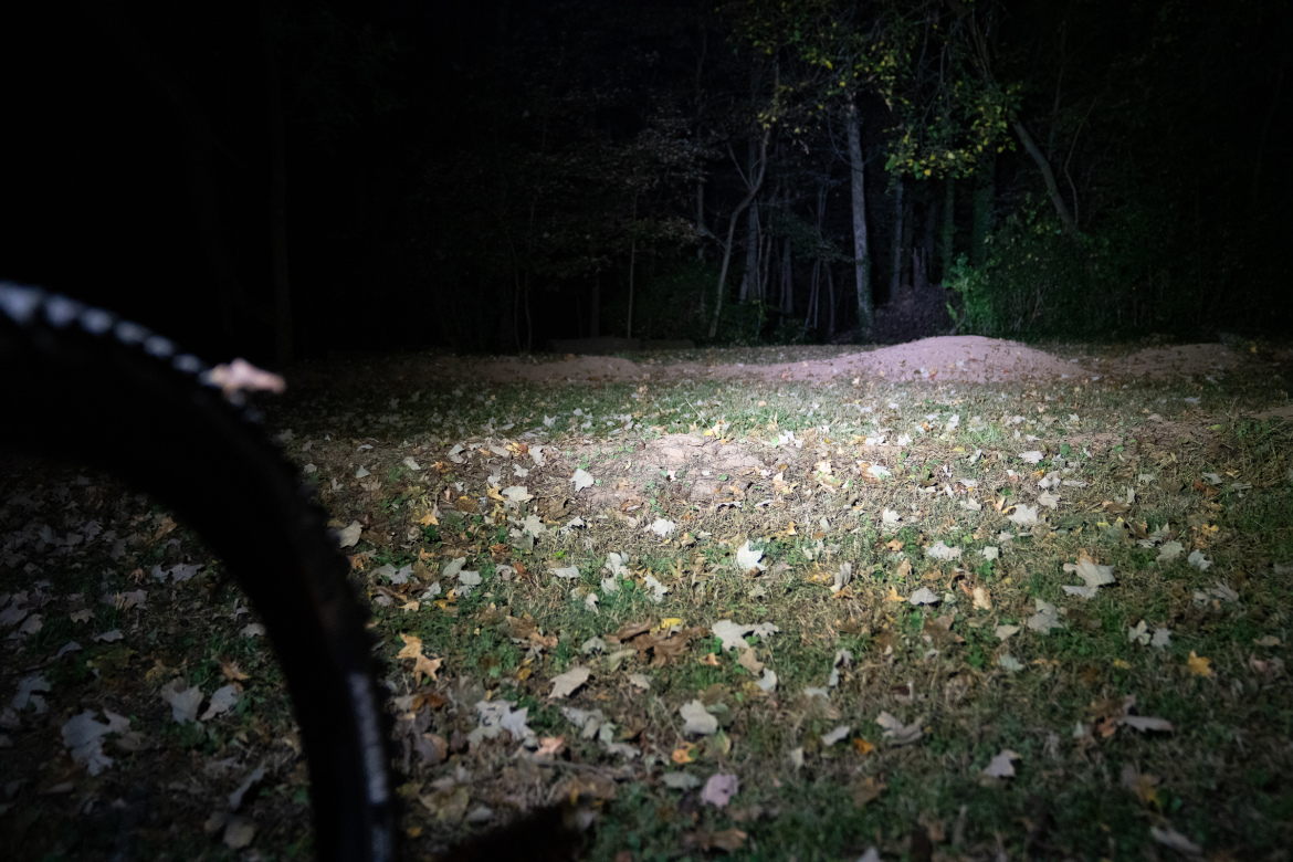 A close-up view of a ground covered with assorted fallen leaves in a dimly lit forest area at night. A portion of a bicycle tire is visible in the foreground, highlighting a trail or path that leads into the darker area of woods.