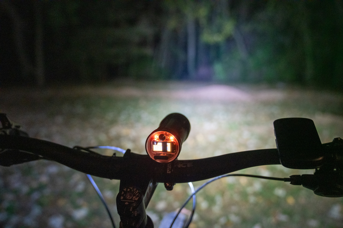 A close-up view of a bicycle handlebar at night, featuring a mounted front light that is illuminated. The light casts a beam onto a path ahead, with a blurred background of trees and scattered leaves on the ground. The scene conveys a sense of nighttime cycling in a natural setting.