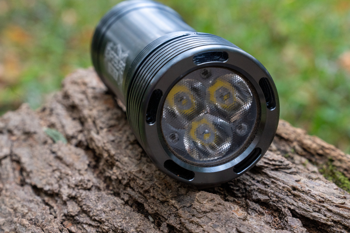 A black flashlight with a textured surface and four bulbs is resting on a piece of tree bark, surrounded by blurred green foliage in a natural outdoor setting.