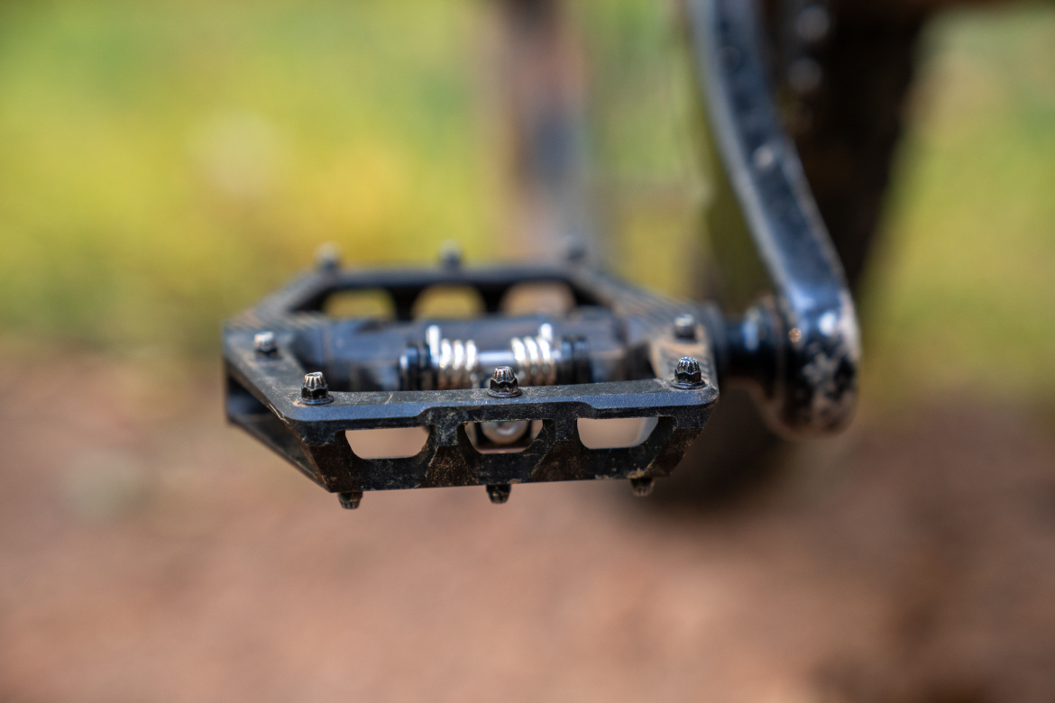 Close-up of a bicycle pedal showing a black metal design with a spring mechanism. The background features a blurred natural setting with greens and browns, indicating an outdoor environment.
