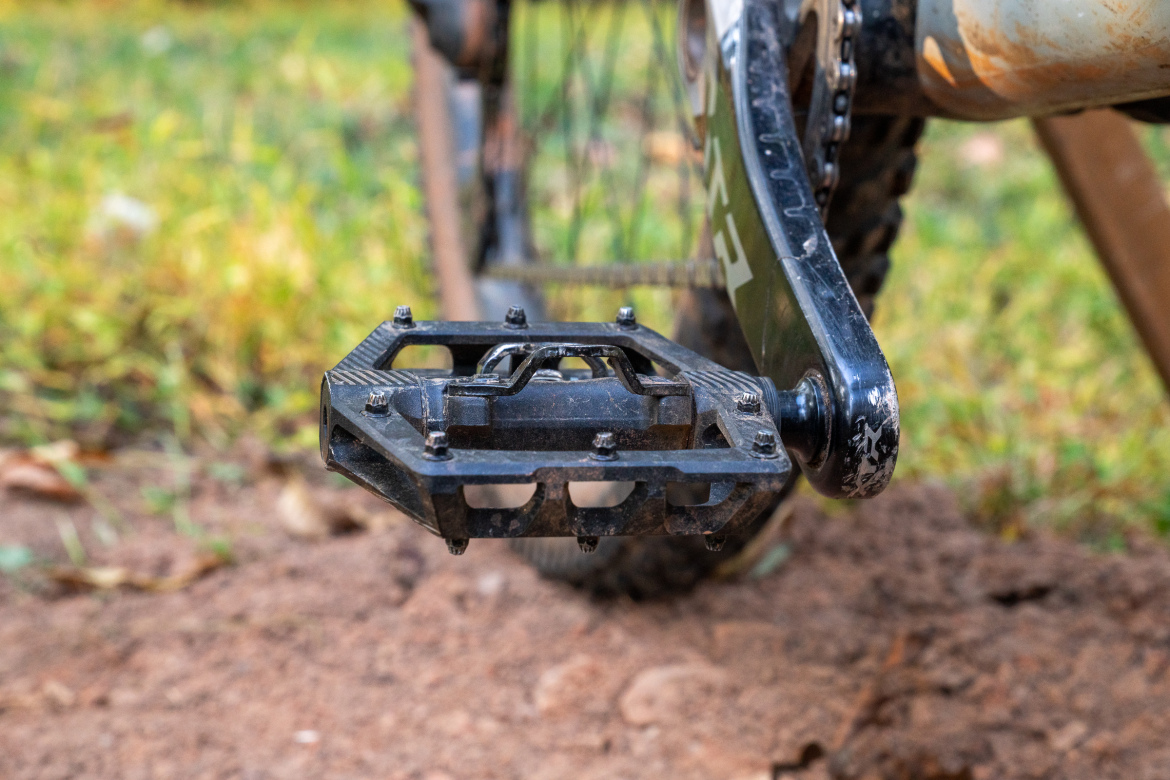 Close-up view of a black bicycle pedal attached to a bike crank, set against a blurred background of grass and dirt. The pedal has a textured surface and metal spikes for grip, indicating it is designed for off-road cycling.
