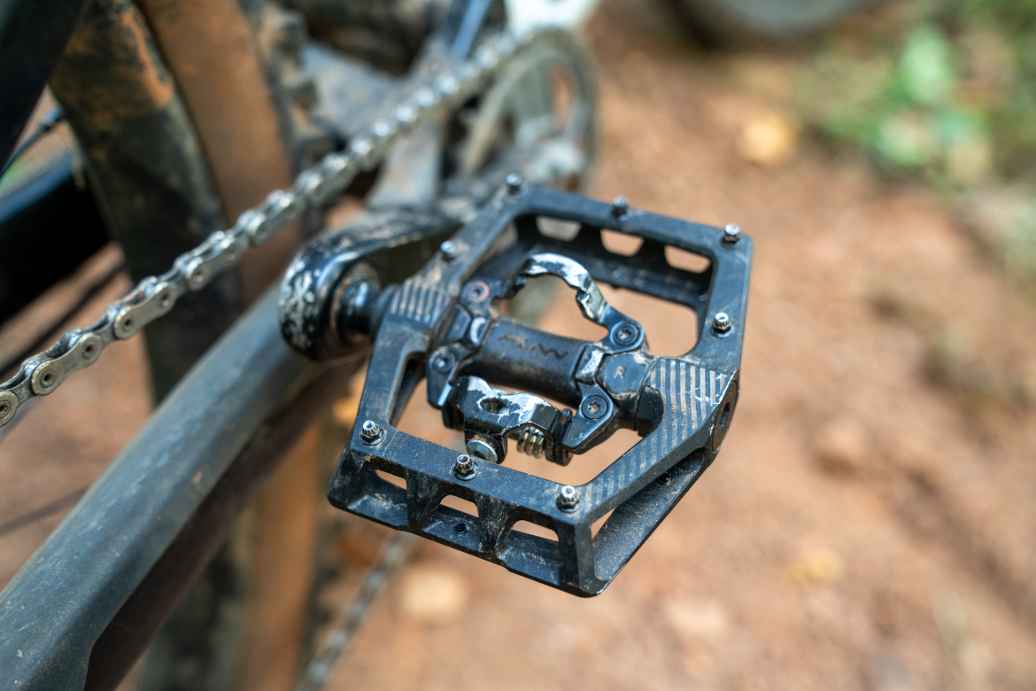 Close-up of a bicycle pedal, showcasing a black, textured surface with visible dirt and wear, attached to a bike chain and crank arm in a natural outdoor setting. The background is blurred, emphasizing the pedal's details and condition.