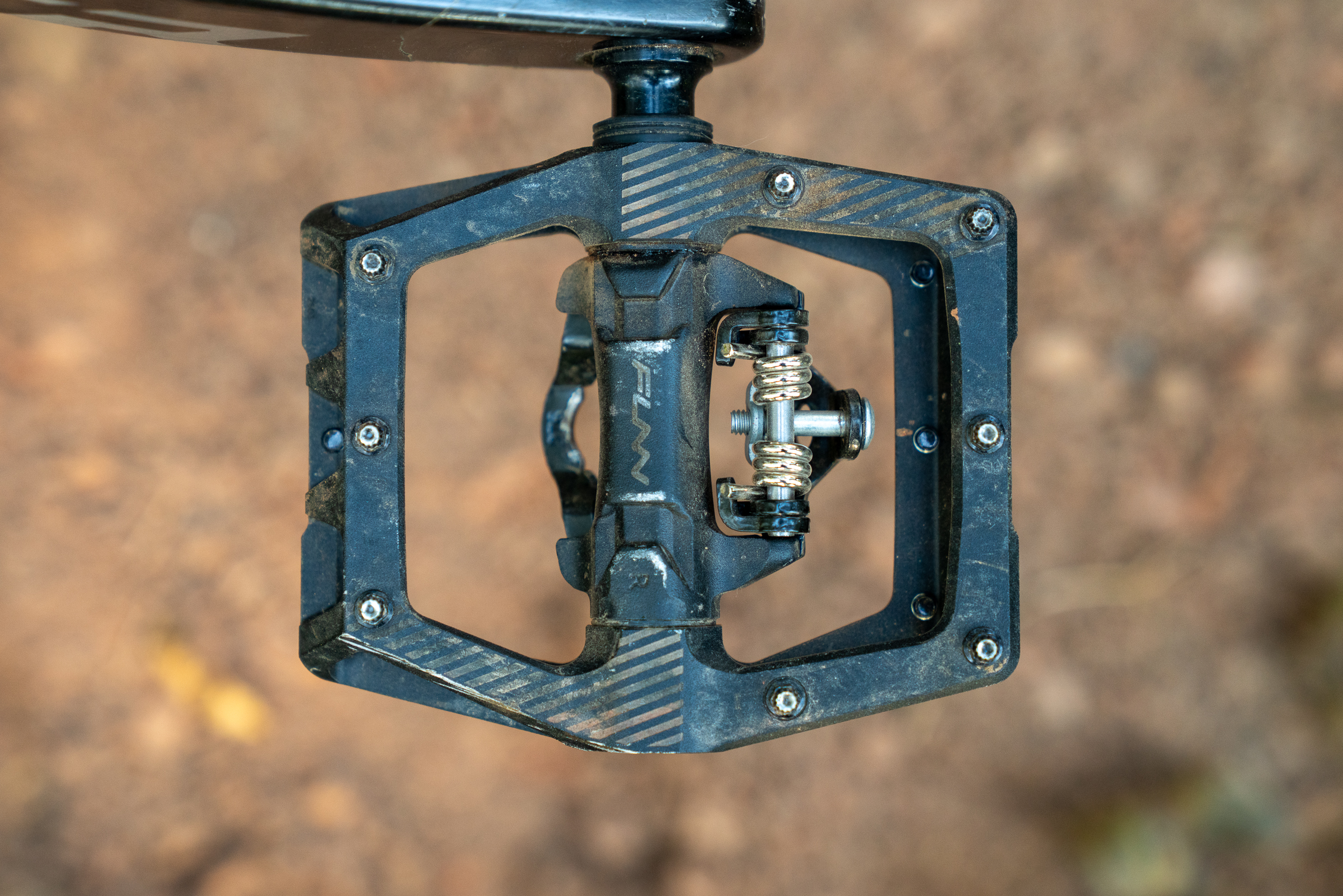 A close-up view of a black bicycle pedal, featuring a platform design with a clip mechanism. The pedal shows signs of use with dirt and scratches, and is positioned against a blurred, earthy background.