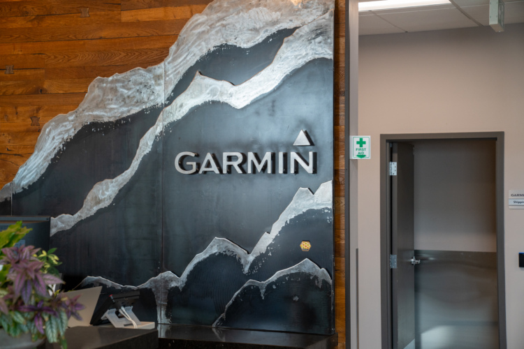 A modern reception area featuring a large, stylized wall art piece with the word "GARMIN" prominently displayed in sleek letters. The background includes wood textures and a design resembling mountain ranges, adding an artistic touch to the corporate environment. A first aid sign is visible to the right of the entrance.