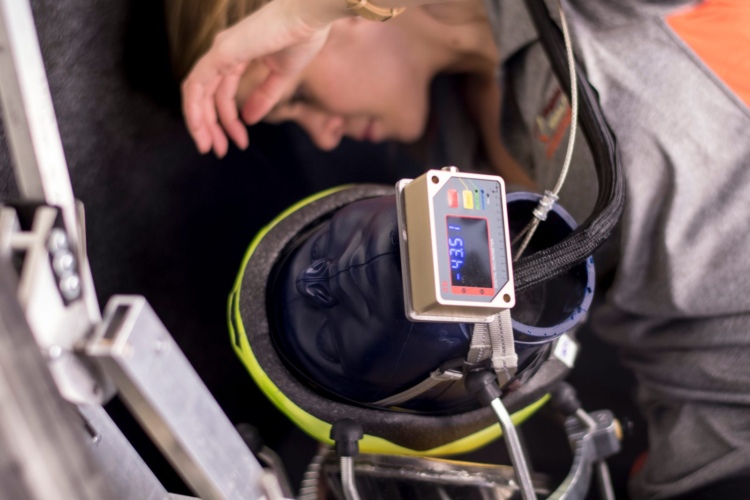 A close-up image featuring a helmet with a digital display attached to it, set against a soft-focused background showing a person bending down. The helmet appears to be designed for safety or testing purposes, indicating the importance of monitoring in the given setting.