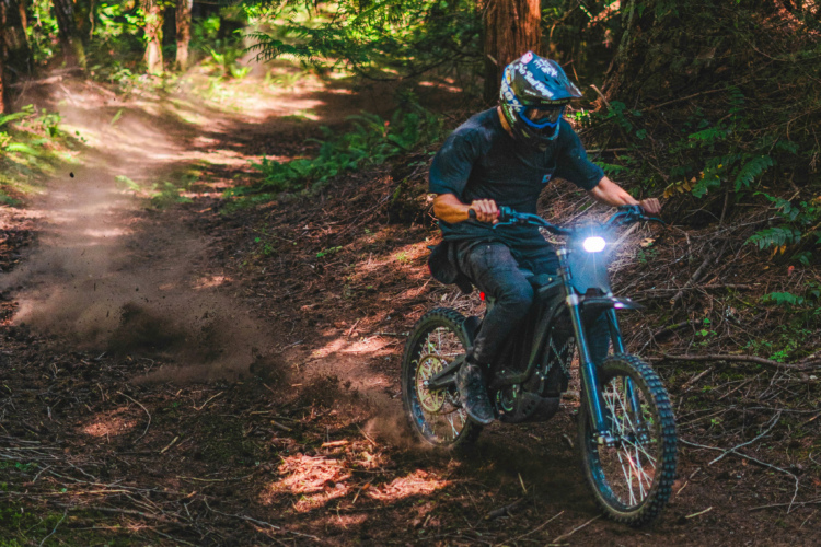 A person wearing a helmet and protective gear rides an electric bike along a dirt trail in a wooded area, kicking up dust behind them as they navigate the path surrounded by trees and foliage.