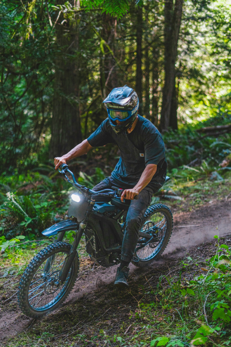 A rider in protective gear maneuvers an electric dirt bike along a narrow trail in a lush, green forest. The bike kicks up dirt as the rider leans forward, navigating the terrain surrounded by tall trees and foliage.