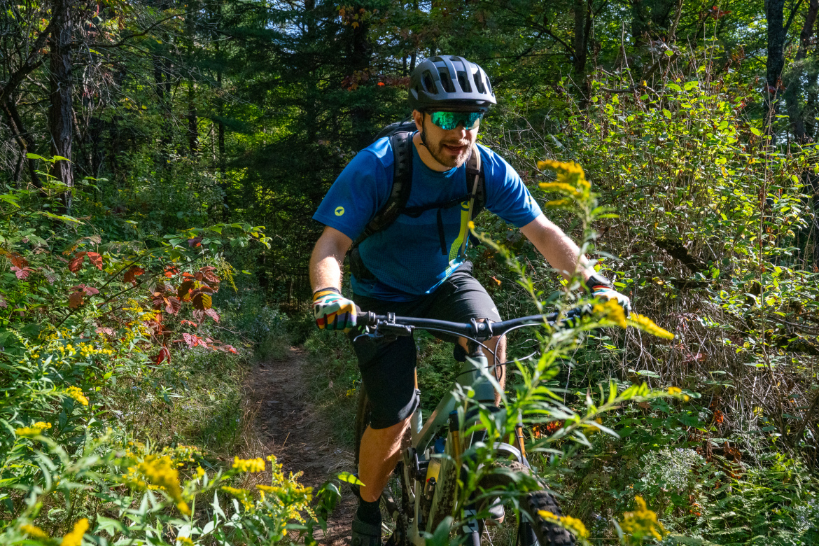 A person riding a mountain bike on a narrow trail surrounded by greenery and colorful foliage, wearing a helmet and sunglasses, with a focus on the handlebars and the path ahead.