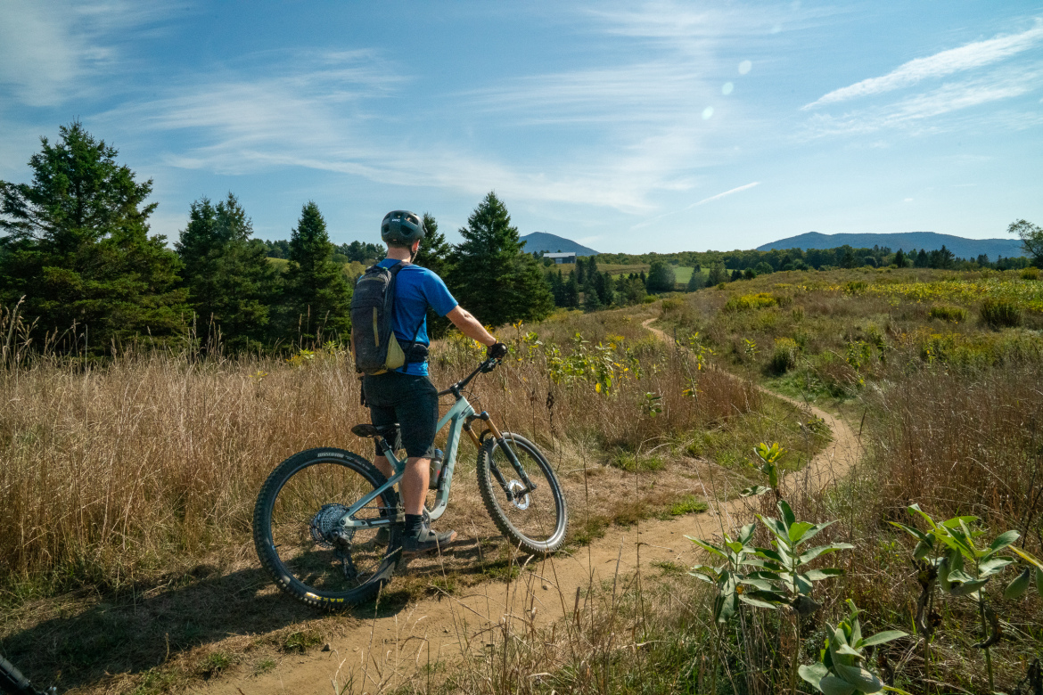 A person riding a mountain bike on a trail through a grassy area, surrounded by trees and distant hills under a clear blue sky. The individual is wearing a helmet and a backpack, gazing down the path ahead.