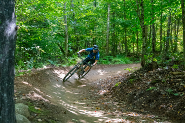 A mountain biker navigates a curved dirt trail through a lush green forest, showcasing dynamic movement and agility while riding. Sunlight filters through the trees, highlighting the vibrant foliage around the path. Black Bear mountain bike trail.