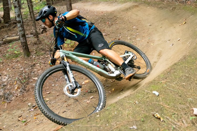 A mountain biker in a blue jersey and black shorts leans into a turn on a dirt trail surrounded by trees, demonstrating agility and control on a green mountain bike.