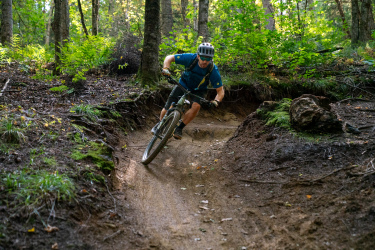 A mountain biker leans into a sharp turn on a dirt trail surrounded by trees and greenery, showcasing skill and balance on rugged terrain. Sidewinder mountain bike trail.