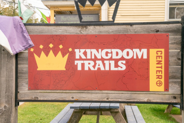 Signage for Kingdom Trails Center, featuring a large yellow crown graphic above the text "KINGDOM TRAILS" in bold white letters on a red background. The bottom right corner includes an arrow symbol indicating direction. A portion of a picnic table is visible in the foreground, with a background of colorful flags and a wooden structure. Kingdom Trails mountain bike trail.