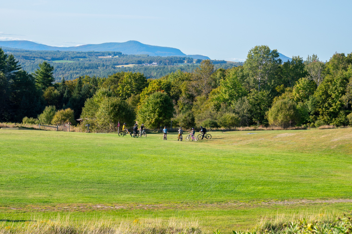 A group of people riding bicycles on a grassy field, surrounded by trees and rolling hills in the background under a clear blue sky.