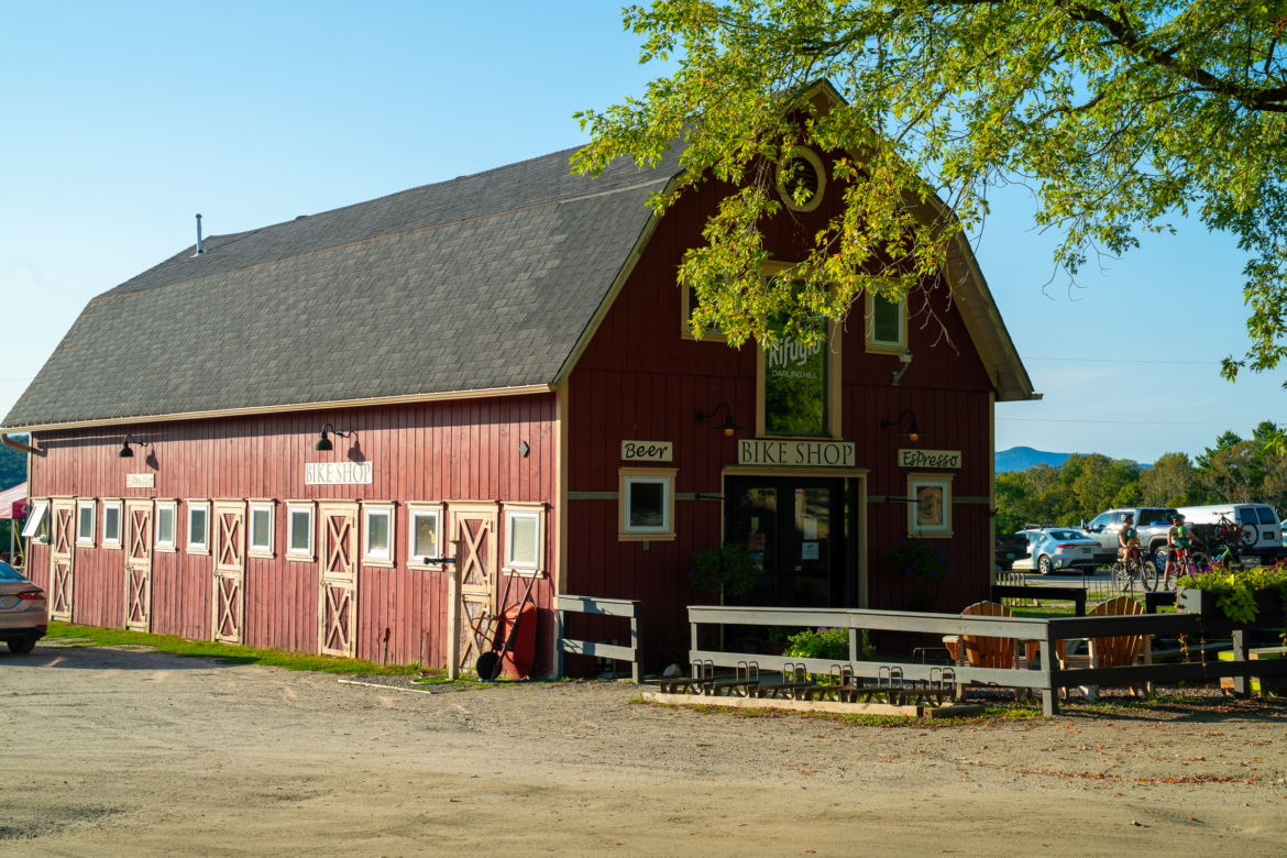 A red barn-style building with a sloped roof, featuring a sign that says "Bike Shop" above the entrance. The facade includes several windows with crossed wooden shutters and additional signs indicating "Beer" and "Espresso." In the foreground, there is a gravel area with parked cars and bike racks, surrounded by greenery and trees under a clear blue sky.