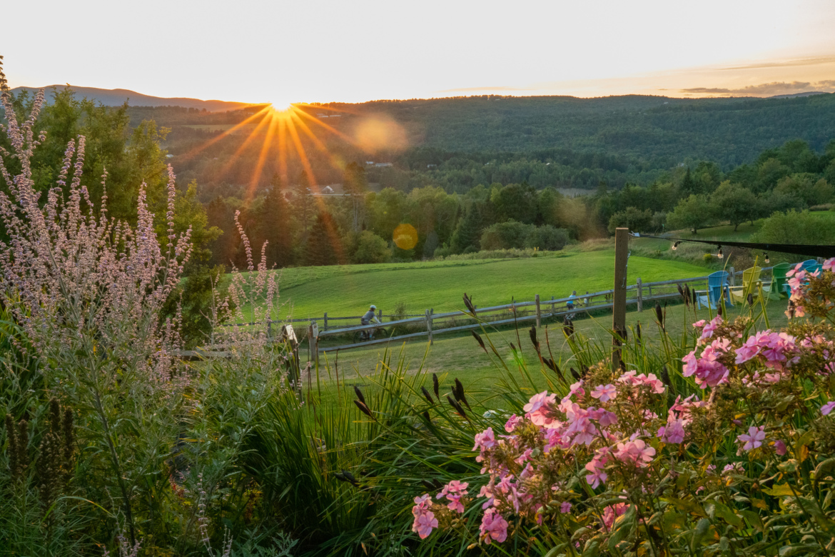 A sunset over a lush green landscape, with vibrant pink flowers in the foreground. The sun's rays create a warm glow as they peek over the distant hills, illuminating the scene. In the background, a few people can be seen enjoying the outdoors near a rustic wooden fence.