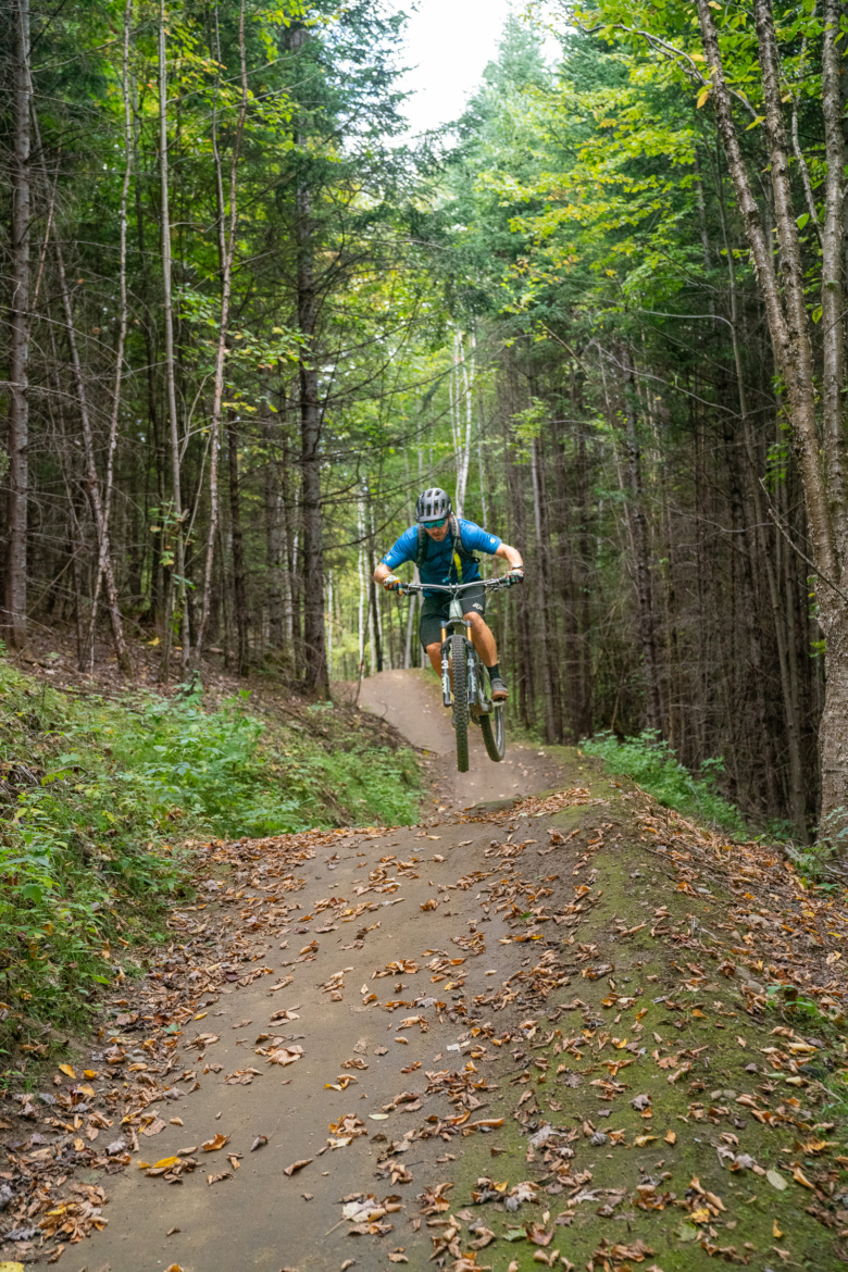 A mountain biker performing a jump on a dirt trail through a dense forest, surrounded by trees in various shades of green and scattered autumn leaves on the ground.