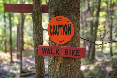 A tree in a wooded area displays two signs: an orange circular sign that reads "CAUTION" and a rectangular red sign below it that says "WALK BIKE." The background features a blurred mix of green foliage, indicating a natural outdoor setting. Widow Maker mountain bike trail.