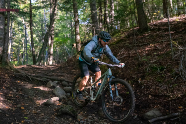 A mountain biker navigating a rocky trail surrounded by trees, captured mid-jump on a forest path. Sunlight filters through the leaves, highlighting the terrain as the rider wears a helmet and protective gear. Widow Maker mountain bike trail.
