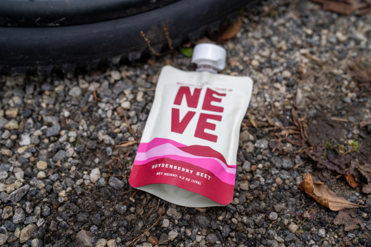 A pouch of NEVE boysenberry beet drink sits on a gravel surface, partially opened with its silver cap on top. The packaging features bold red and pink graphics on a cream background, prominently displaying the product name and flavor. In the background, a black tire is partially visible, with some leaves scattered around in the vicinity.