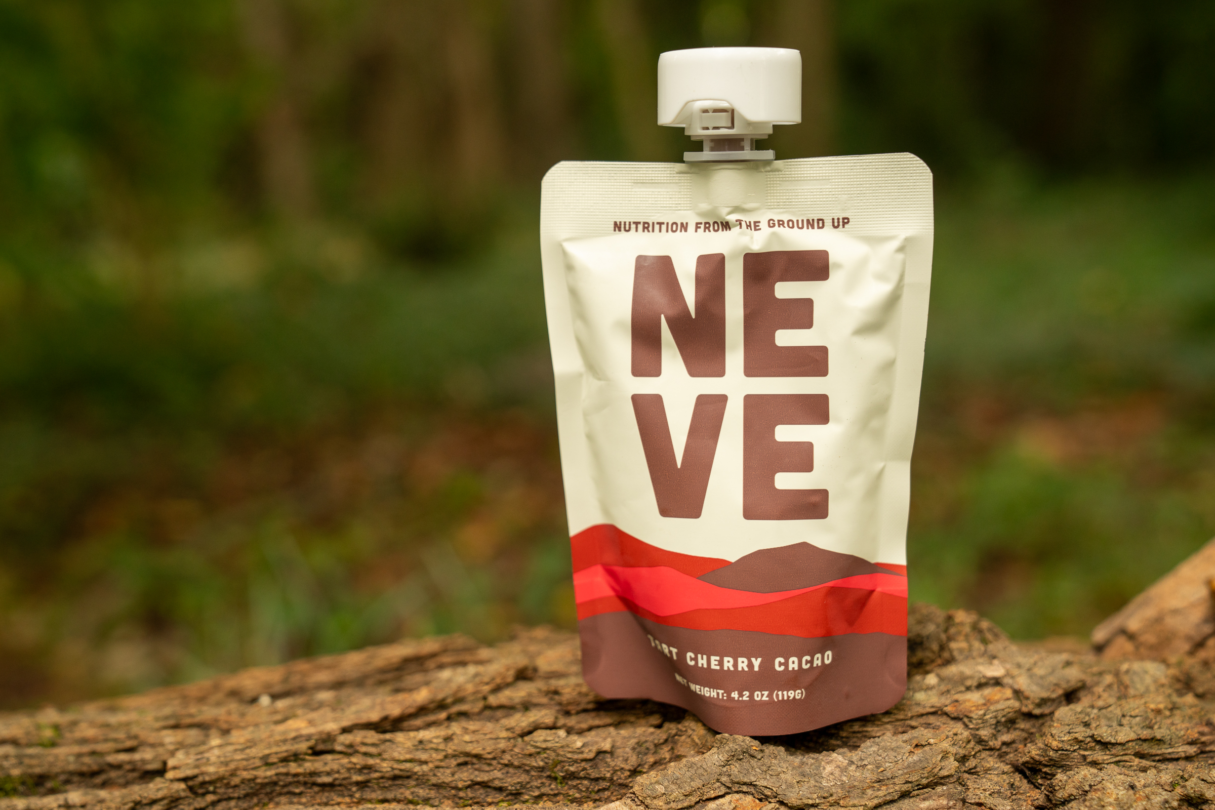 A pouch of NEVE nutritional supplement is displayed on a log, featuring the flavor 'Mont Cherry Cacao.' The packaging has a white background with bold brown lettering and a stylized design in red and brown at the bottom. The setting includes a blurred natural background with greenery.