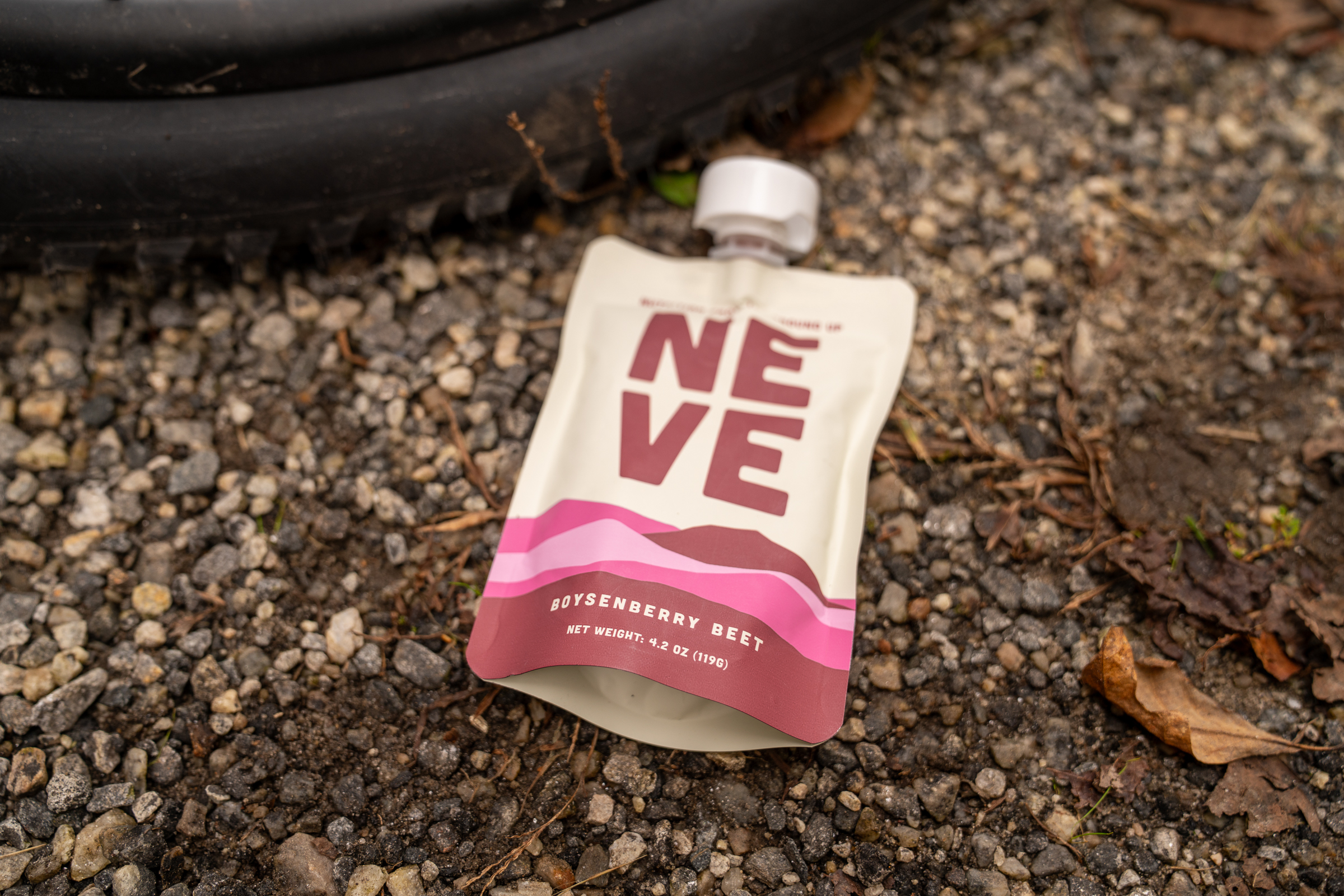 A close-up of a beige pouch labeled "NEVE" featuring the flavor "Boysenberry Beet." The pouch is partially open and placed on a gravel surface with small stones and scattered leaves in the background. A dark tire is visible in the upper part of the image.