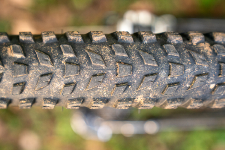 Close-up view of a bicycle tire tread, showing a textured surface with distinct patterns designed for traction. The tire has visible dirt and debris, indicating it has been used on rugged terrain. The background is slightly blurred, suggesting an outdoor setting.