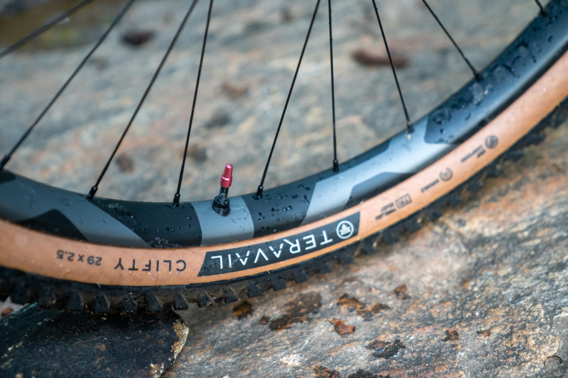 Close-up of a mountain bike tire with a brown rubber tread, showing water droplets on the surface. The tire has a black and grey colored rim with visible branding, and a red valve stem is prominently displayed. The background features a rocky surface, suggesting an outdoor setting.