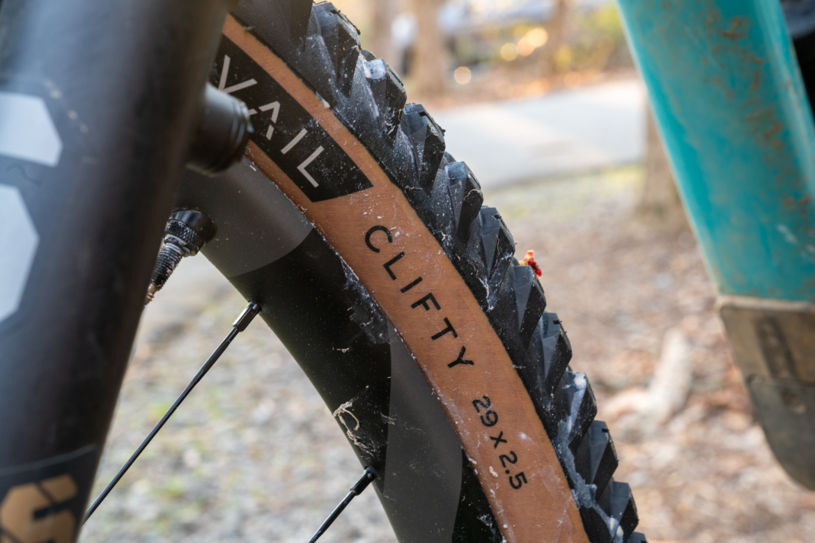 Close-up view of a mountain bike tire featuring knobby tread and the brand name "Clifty" with specifications "29x2.5" on the sidewall. The tire is mounted on a black wheel, showcasing a portion of the bike's frame and spokes. The background includes a blurred natural setting with trees and dirt path.