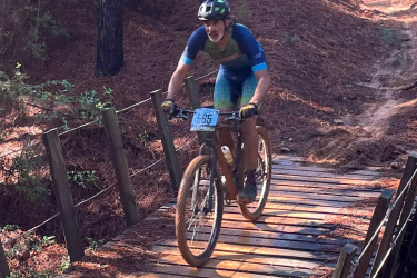 A cyclist in a blue and green kit riding a mountain bike across a wooden bridge in a forested area, surrounded by pine needles and trees. The cyclist is wearing a helmet and appears to be focused on navigating the trail. Rocky Hill Ranch mountain bike trail.