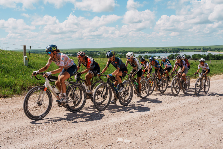A group of cyclists riding in a line on a gravel road, surrounded by lush green fields and a blue sky with fluffy clouds. The cyclists are wearing colorful jerseys and helmets, focused on the race. A scenic landscape with trees and a body of water is visible in the background.