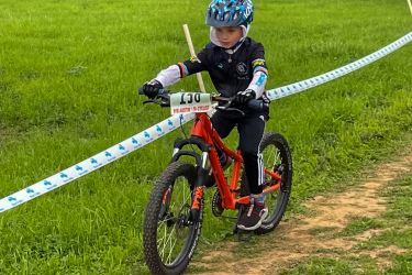 A young child riding a bright orange mountain bike on a dirt path surrounded by green grass, wearing a blue helmet and cycling gear. The path is marked with blue and white tape, indicating a cycling event. Rocky Hill Ranch mountain bike trail.