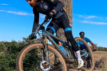 A mountain biker in a black and blue cycling outfit navigates a dirt trail, leaning forward as he rides over a small incline. Another cyclist follows behind on a separate bike. The background features a clear blue sky and greenery along the trail. Rocky Hill Ranch mountain bike trail.